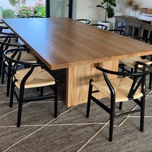 May include: A large wooden dining table with a natural wood finish and black chairs with woven seats. The table is set in a dining room with a rug on the floor.
