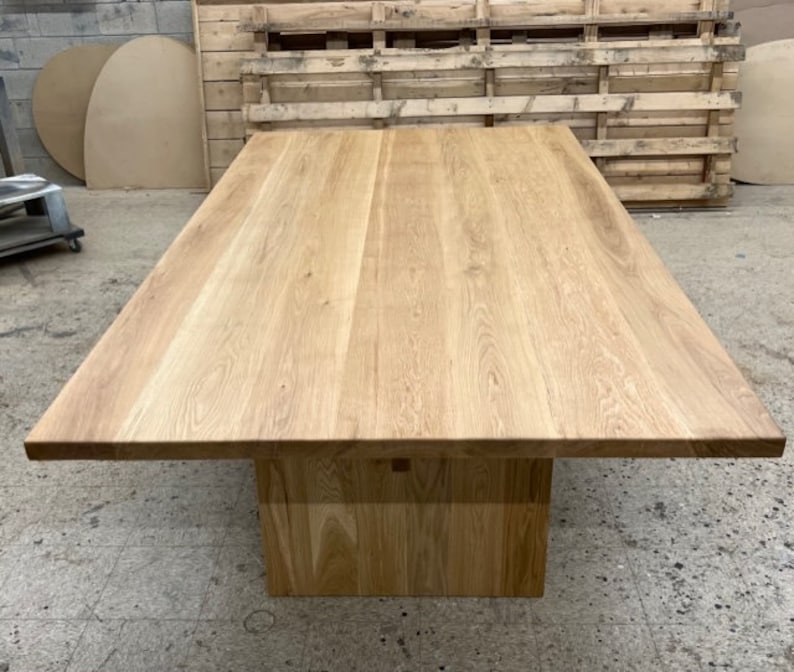 May include: A large, rectangular, wooden dining table with a natural wood finish. The table has a thick, solid top and a simple, square base.