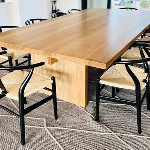 May include: A large wooden dining table with a natural wood finish and a set of black chairs with woven cane seats. The table is set in a modern dining room with a patterned rug.