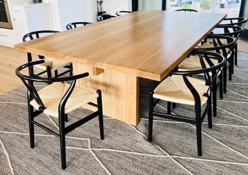 May include: A large wooden dining table with a natural wood finish and a set of black chairs with woven cane seats. The table is set in a modern dining room with a patterned rug.