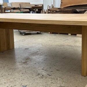 May include: A long, rectangular wooden table with a natural finish. The table has a simple design with a wide, flat top and a single, wide, rectangular base.