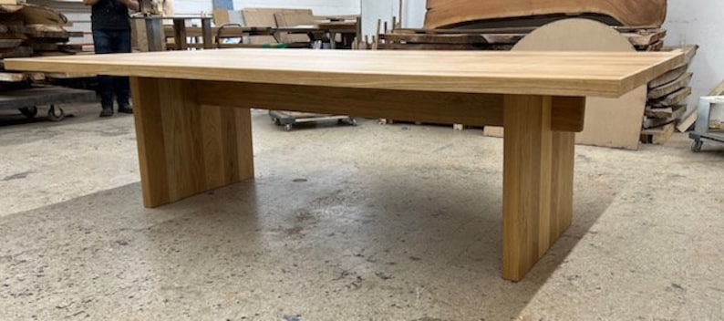 May include: A long, rectangular wooden table with a natural finish. The table has a simple design with a wide, flat top and a single, wide, rectangular base.