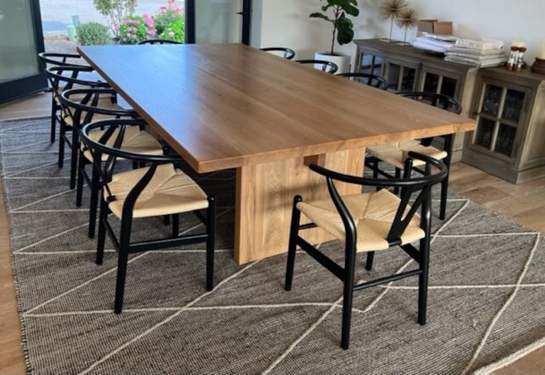 May include: A large wooden dining table with a natural wood finish and black chairs with woven seats. The table is set in a dining room with a rug on the floor.