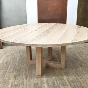 May include: A round wooden dining table with a natural wood finish and a simple, modern design. The table has a square base with four legs.