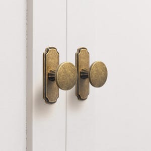 Może przedstawiać: Zbliżenie dwóch antycznych mosiężnych gałek do szafek o okrągłym kształcie. Gałki są zamontowane na białych drzwiach szafki, prezentując klasyczny styl okuć. Gałki mają prostokątną płytkę tylną.