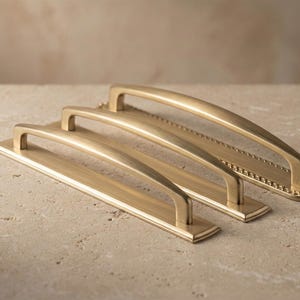 May include: Three gold-coloured cabinet pulls of varying sizes are arranged on a light-coloured surface. Each pull features a rectangular base with a curved handle. The largest pull has a decorative edge.