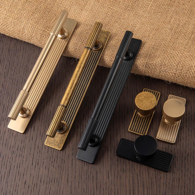 Puede incluir: Un conjunto de herrajes para armarios en acabados dorado, lat&oacute;n antiguo y negro. El hardware incluye tiradores largos y pomos redondos, todos con un dise&ntilde;o acanalado. Los art&iacute;culos est&aacute;n dispuestos sobre una superficie de madera oscura.