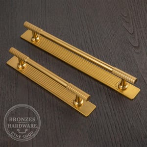 Puede incluir: Dos tiradores dorados con dise&ntilde;o acanalado. Cada tirador tiene una base rectangular y un mango largo y cil&iacute;ndrico. Los tiradores se muestran sobre una superficie de madera oscura. El logotipo de la tienda Etsy es visible.