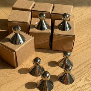 May include: Silver-colored bottle stoppers with a spherical top and a conical base are displayed. Some stoppers are on top of small, square, brown cardboard boxes. The stoppers are arranged on a light-colored wooden surface.