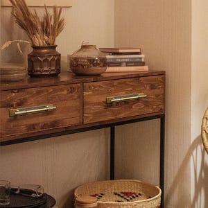 Puede incluir: Una consola de madera con dos cajones, con tiradores de color lat&oacute;n. La mesa est&aacute; adornada con objetos decorativos, incluyendo un jarr&oacute;n con plantas secas, un tarro de cristal y una pila de libros. Debajo hay una cesta tejida.