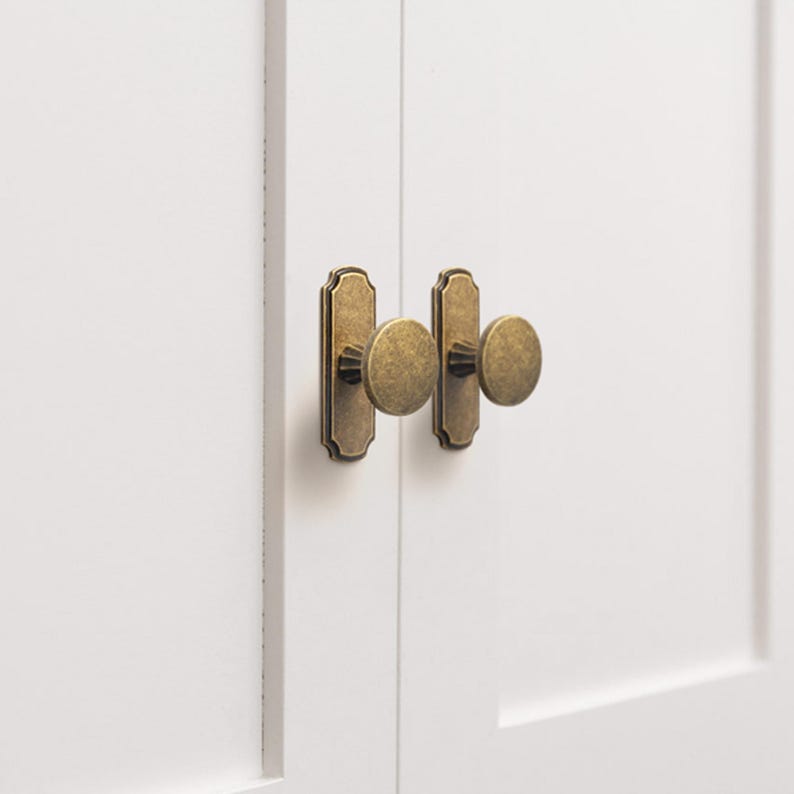 May include: Close-up of two cabinet doors with brass-colored hardware. Each door features a rectangular backplate with rounded edges and a circular knob. The hardware has an antique finish, contrasting with the clean, white surface of the cabinet doors. The doors are positioned side by side.