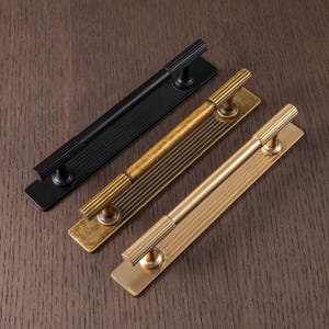 Puede incluir: Tres tiradores de puerta met&aacute;licos en negro, lat&oacute;n antiguo y dorado. Cada tirador tiene una placa trasera rectangular con ranuras verticales y un asa cil&iacute;ndrica con crestas horizontales. El herraje est&aacute; dise&ntilde;ado para armarios o puertas.