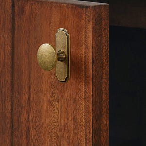May include: Close-up of a wooden cabinet door with a brass knob and backplate. The door is a rich, warm brown with visible wood grain. The hardware has an aged, antique finish, adding a touch of vintage charm to the cabinet.