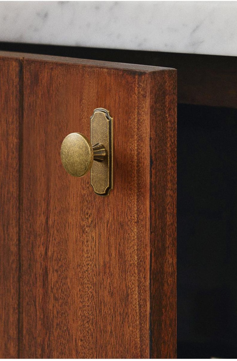 May include: Close-up of a wooden cabinet door with a brass knob and backplate. The door is a rich, warm brown with visible wood grain. The hardware has an aged, antique finish, adding a touch of vintage charm to the cabinet.