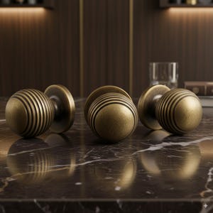 May include: Three antique brass door knobs with a round, grooved design. The knobs are set against a dark, marble-like surface, with a blurred background. The knobs have a vintage, aged appearance.