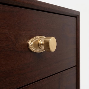 May include: A close-up of a dark brown wooden drawer with a gold-colored knob. The knob has a round, ridged handle and an oval backplate with a beaded edge. The drawer is part of a larger piece of furniture.