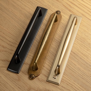 May include: Three cabinet pulls in black, antique brass, and brushed gold finishes. Each pull features a rectangular backplate and a curved handle. The hardware is displayed on a light wood surface.