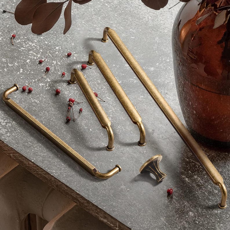 May include: A collection of antique brass cabinet hardware, including several pull handles and a knob. The hardware has a warm, aged finish and a classic design. The pieces are arranged on a gray surface.