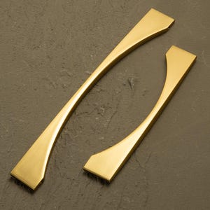 May include: Two gold-toned cabinet pulls with a modern design. One pull is long and curved, while the other is shorter with a similar shape. The pulls have a matte finish and are set against a dark, textured background.