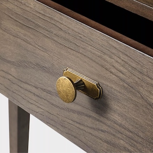 May include: Close-up of a wooden drawer with a brass knob and decorative backplate. The drawer is made of gray wood with visible grain. The knob is round and the backplate is rectangular with a curved edge.