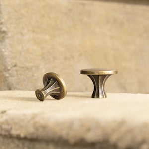 May include: Two antique brass cabinet knobs. The knobs have a round top and a decorative, fluted base. One knob is upright, and the other is lying on its side. The knobs are set against a neutral background.