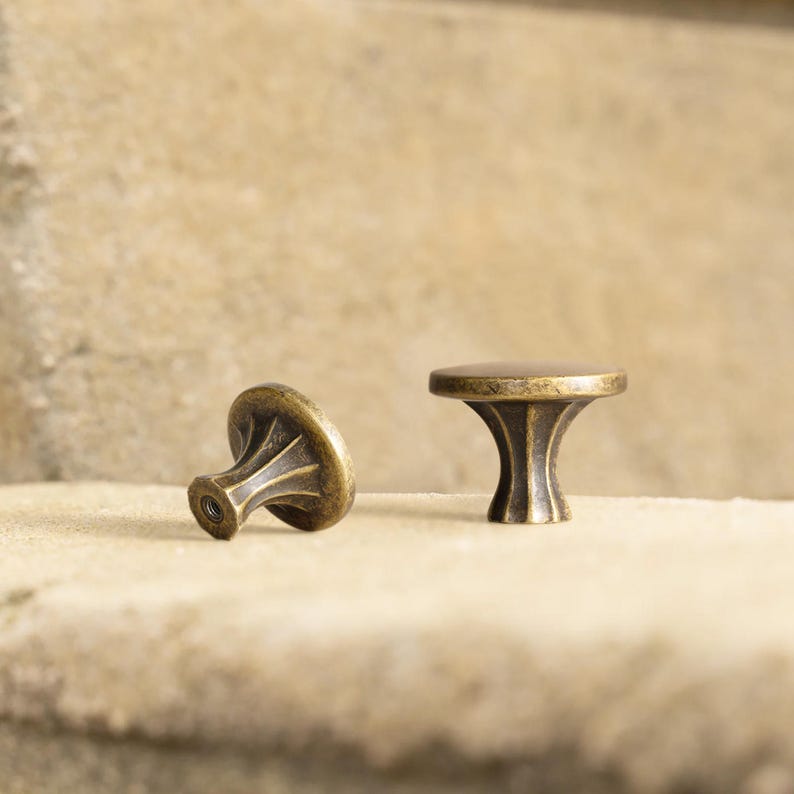 May include: Two antique brass cabinet knobs. The knobs have a round top and a decorative, fluted base. One knob is upright, and the other is lying on its side. The knobs are set against a neutral background.