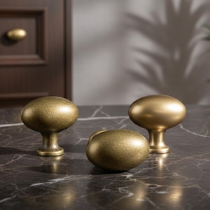 May include: Three antique brass cabinet knobs with an oval design. One knob is lying down, while the other two are upright. The knobs have a textured finish and are set against a dark marble surface.