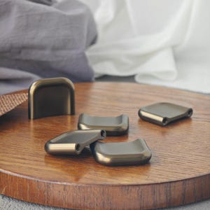 Könnte beinhalten: Bronzefarbene Schrankgriffe und -knöpfe in verschiedenen Formen. Die Hardware ist auf einer Holzoberfläche angeordnet, mit einem neutralen Stoffhintergrund. Die Griffe haben ein gebogenes Design, und einige haben vorgebohrte Löcher für Schrauben.