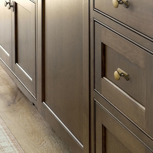 May include: Close-up of dark brown kitchen cabinets with brass hardware. The cabinets have a simple, classic design with framed doors and drawers. The wood grain is visible, adding texture. The floor is light-colored wood.
