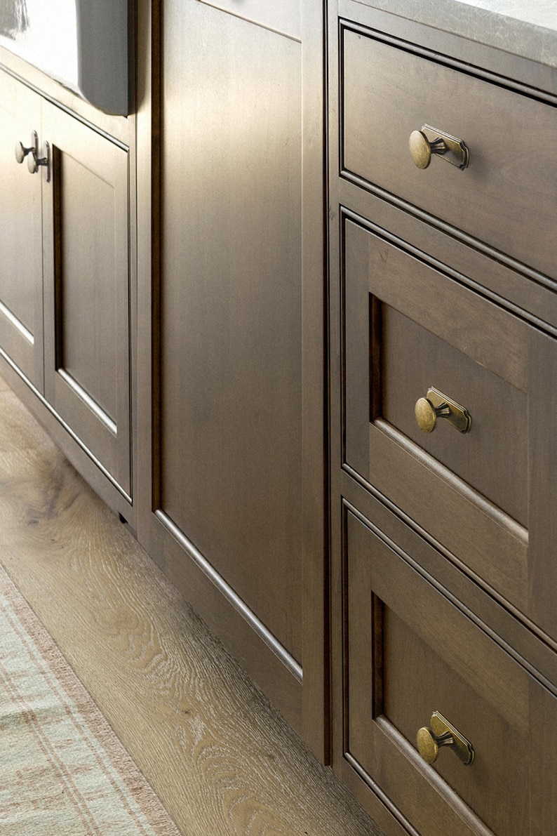 May include: Close-up of dark brown kitchen cabinets with brass hardware. The cabinets have a simple, classic design with framed doors and drawers. The wood grain is visible, adding texture. The floor is light-colored wood.