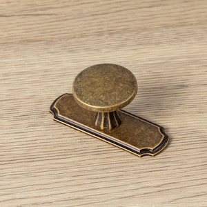 May include: Antique brass cabinet pull with a round knob and rectangular backplate. The knob is circular and sits atop a decorative base. The backplate has a decorative edge. This hardware is suitable for cabinets and drawers.