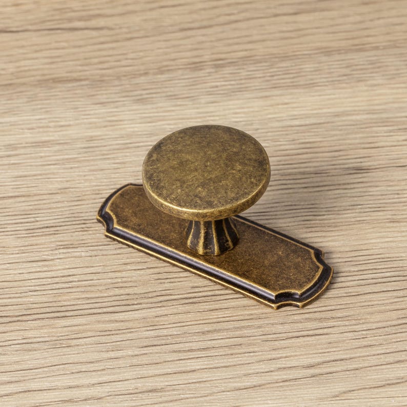 May include: Antique brass cabinet pull with a round knob and rectangular backplate. The knob is circular and sits atop a decorative base. The backplate has a decorative edge. This hardware is suitable for cabinets and drawers.