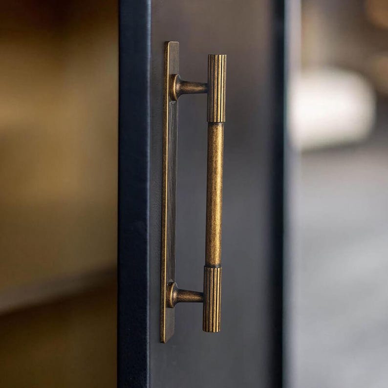 Puede incluir: Primer plano de un tirador de armario de lat&oacute;n. El tirador tiene una placa trasera rectangular y una barra vertical con un dise&ntilde;o acanalado. El acabado es de color oro antiguo c&aacute;lido, y el tirador est&aacute; montado en una puerta de armario azul oscuro.
