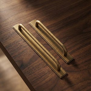 Op de afbeelding: Twee antieke messing kastgrepen met een rechthoekige achterplaat en een gebogen handvat. De grepen hebben een warme goudkleur en zijn geplaatst op een donkere houten ondergrond. Ideaal voor een vintage touch.