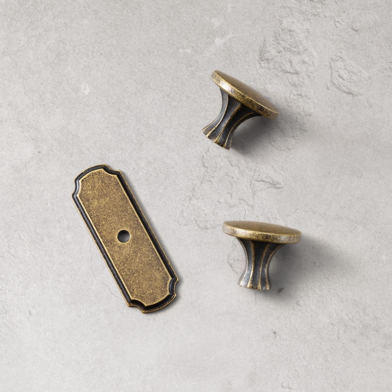 May include: An overhead shot of three antique brass cabinet hardware pieces. There are two mushroom-shaped knobs and one rectangular backplate with a single hole. The hardware has a distressed finish with dark accents.