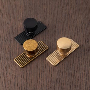 Puede incluir: Tres tiradores de armario en diferentes acabados: negro, lat&oacute;n y dorado. Cada tirador tiene una parte superior cil&iacute;ndrica con ranuras verticales, montada sobre una placa trasera rectangular con ranuras paralelas. Los tiradores se muestran sobre una superficie de madera oscura.