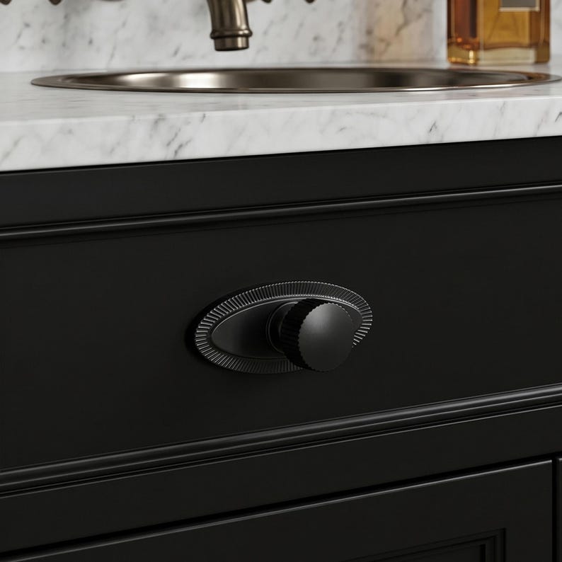 May include: Close-up of a black cabinet drawer with a decorative oval knob. The knob has a ridged edge and a round, textured center. The cabinet is part of a bathroom vanity with a marble countertop and a metal faucet.