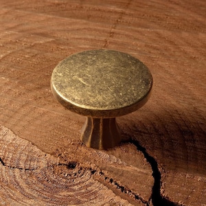 May include: A round, antique brass cabinet knob with a slightly domed top and a decorative, fluted base. The knob is set against a textured, brown wooden surface, creating a rustic aesthetic. This hardware is suitable for drawers and cabinets.