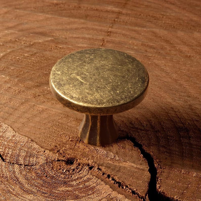 May include: A round, antique brass cabinet knob with a slightly domed top and a decorative, fluted base. The knob is set against a textured, brown wooden surface, creating a rustic aesthetic. This hardware is suitable for drawers and cabinets.