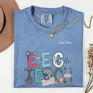 May include: A blue crew neck t-shirt featuring a graphic design with the text EEG TECH. The letters are decorated with medical equipment illustrations including electrodes, a syringe, and a brain wave monitor. The shirt has a Comfort Colors label.