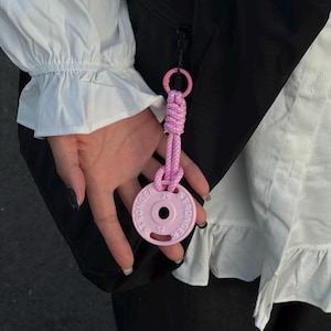 Può includere: Un portachiavi a forma di piastra rosa con la scritta "STRONGER" stampata sul bordo. Il portachiavi ha una corda e un anello rosa. La piastra ha il numero "25" stampato.