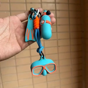以下が含まれることがあります： 3つのミニチュアスキューバダイビングアクセサリーが付いたキーホルダー。オレンジ色のアクセントが入った水色のスイムフィン、オレンジ色のストラップが付いた水色の酸素ボンベ、オレンジ色のフレームが付いた水色のダイビングマスクが、青いコードで繋がれています。