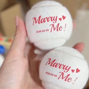 Puede incluir: Dos pelotas de tenis blancas con las palabras "Marry Me!" impresas en rojo, junto con la fecha "2026-02-09". El texto está acompañado de dos pequeños corazones rojos. Las pelotas están en una mano.