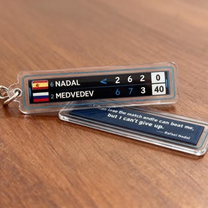 May include: A clear acrylic keychain featuring a tennis scoreboard with the names Nadal and Medvedev, and a quote by Rafael Nadal. The scoreboard displays the match scores. The keychain has a silver chain.