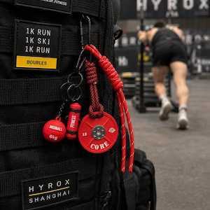 May include: A black tactical backpack featuring Hyrox branding patches and a series of miniature fitness equipment keychains. The attachments include a red 6kg medball, a red fitness cylinder, and a red weight plate labeled 15 and CORE.