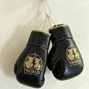 Può includere: Un paio di guantoni da boxe neri con bordi dorati e cuciture bianche, appesi a un cordino bianco. Ogni guantone presenta una grafica dorata di due tori muscolosi e la scritta "TWORUS B&T MUAY THAI" stampata sulla superficie in pelle.