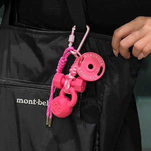 Op de afbeelding: Een roze sleutelhanger met miniatuur gymuitrusting, waaronder een halter, een kettlebell en een gewichtsschijf. Op de gewichtsschijf staat het woord "STRONGER". De sleutelhanger is bevestigd aan een zwarte tas met een zilveren karabijnhaak.