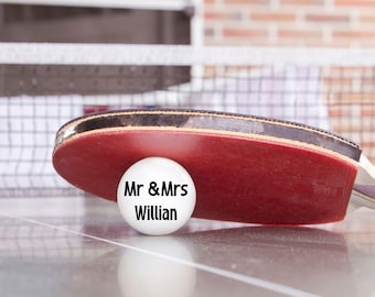May include: A red ping pong paddle with a black handle rests on a table next to a white ping pong ball. The ball has the text "Mr & Mrs Willian" printed on it. The background includes a net and a brick wall.