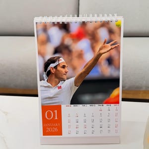 May include: A white desk calendar featuring a tennis player in action, with a tennis ball in the air. The calendar shows the month of January 2026. The calendar is on a white surface.