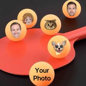 May include: Orange ping pong balls with faces and a red paddle. The balls feature various faces, including a cat, a dog wearing sunglasses, and human faces. One ball has the text "Your Photo".
